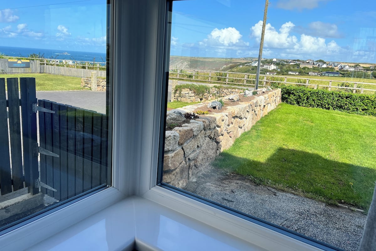 A view from a window overlooking a grassy area with a stone wall, a wooden fence, and a distant coastline.