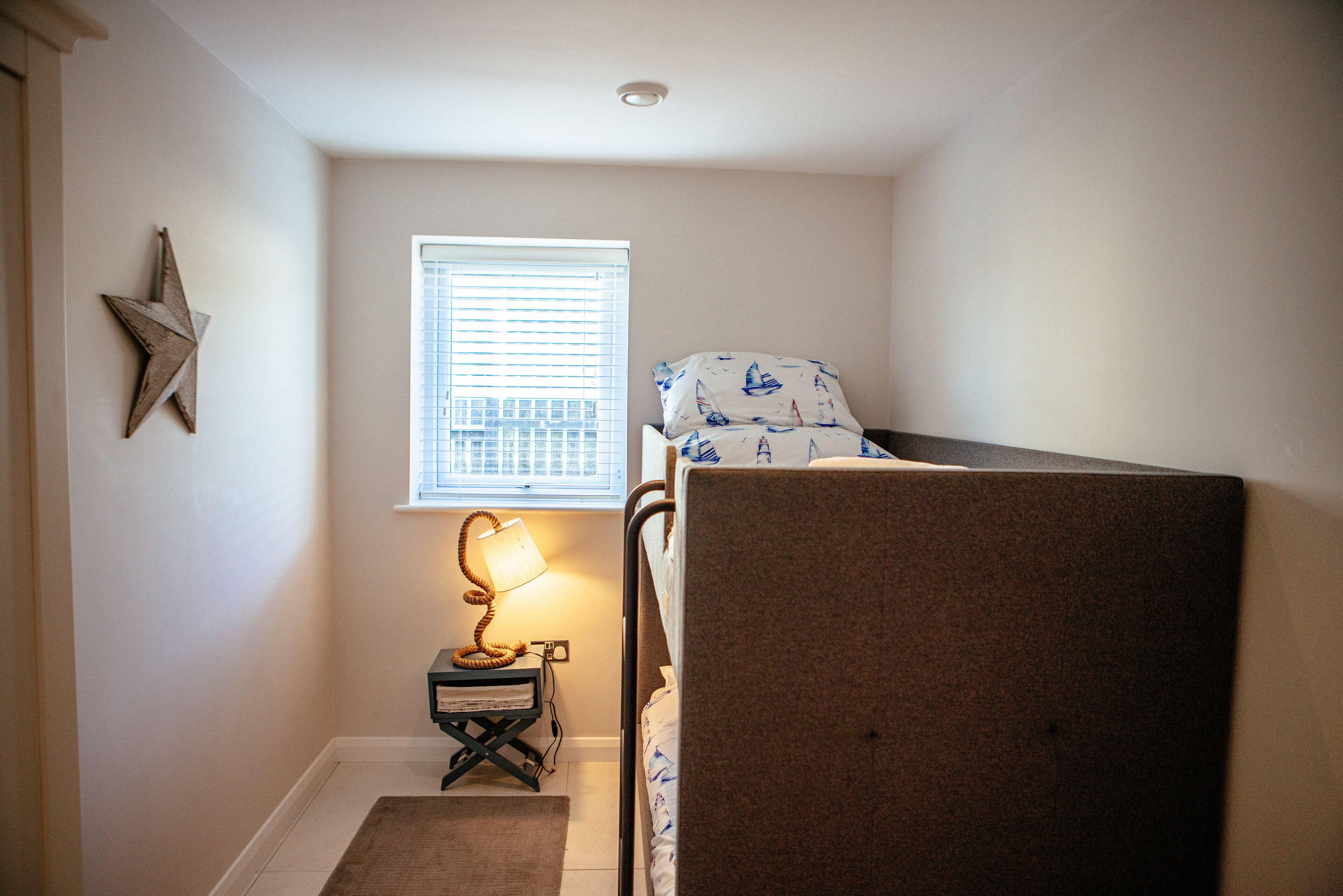 A small, neatly arranged bedroom with a bunk bed, a bedside lamp, a star decoration, and a window with blinds.