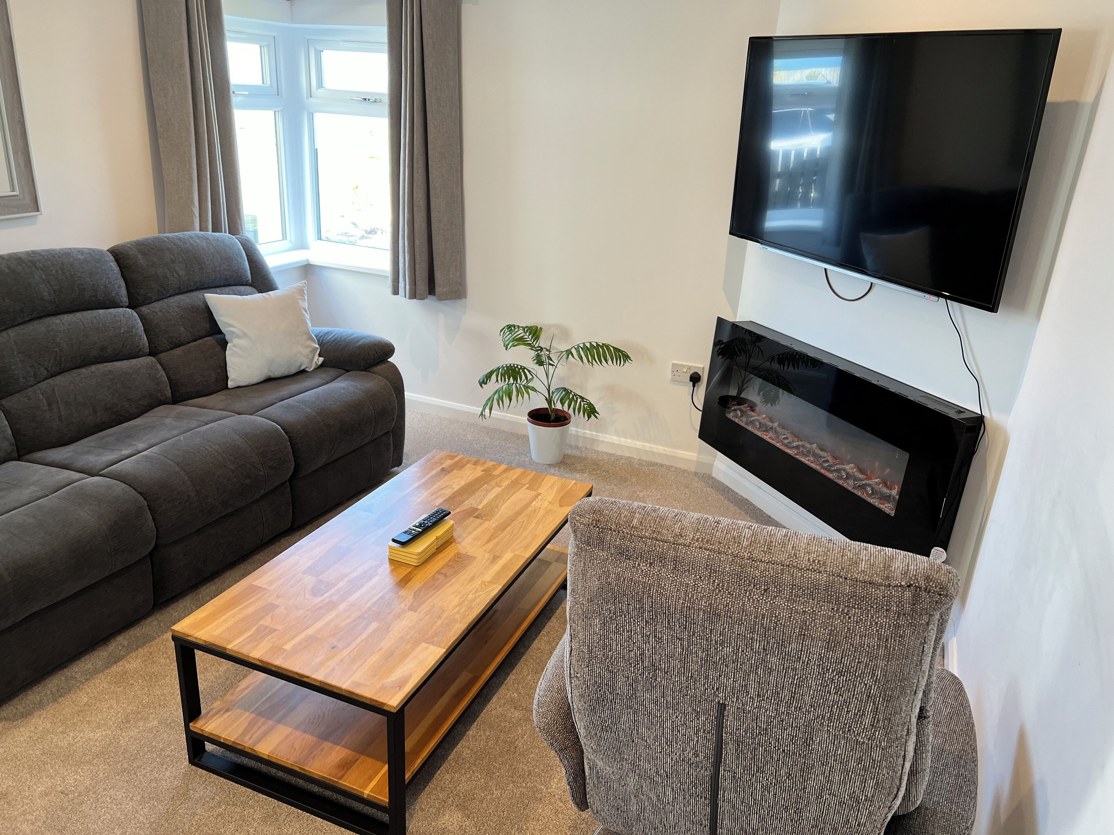 A living room with a dark sofa, a wooden coffee table, a television mounted on a wall, and a potted plant.