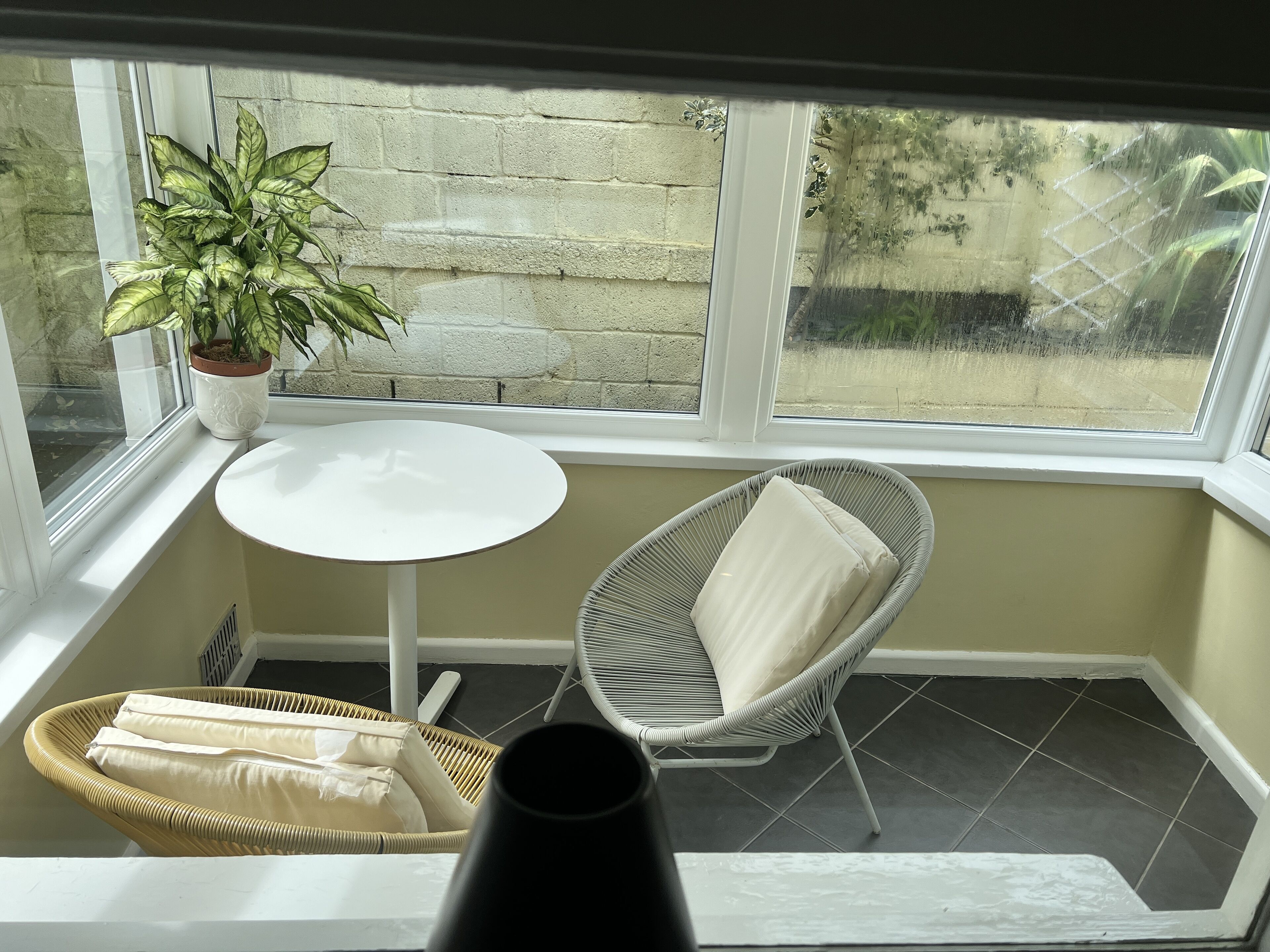 Habitación de esquina con una mesa blanca, una silla de mimbre y una planta en maceta.