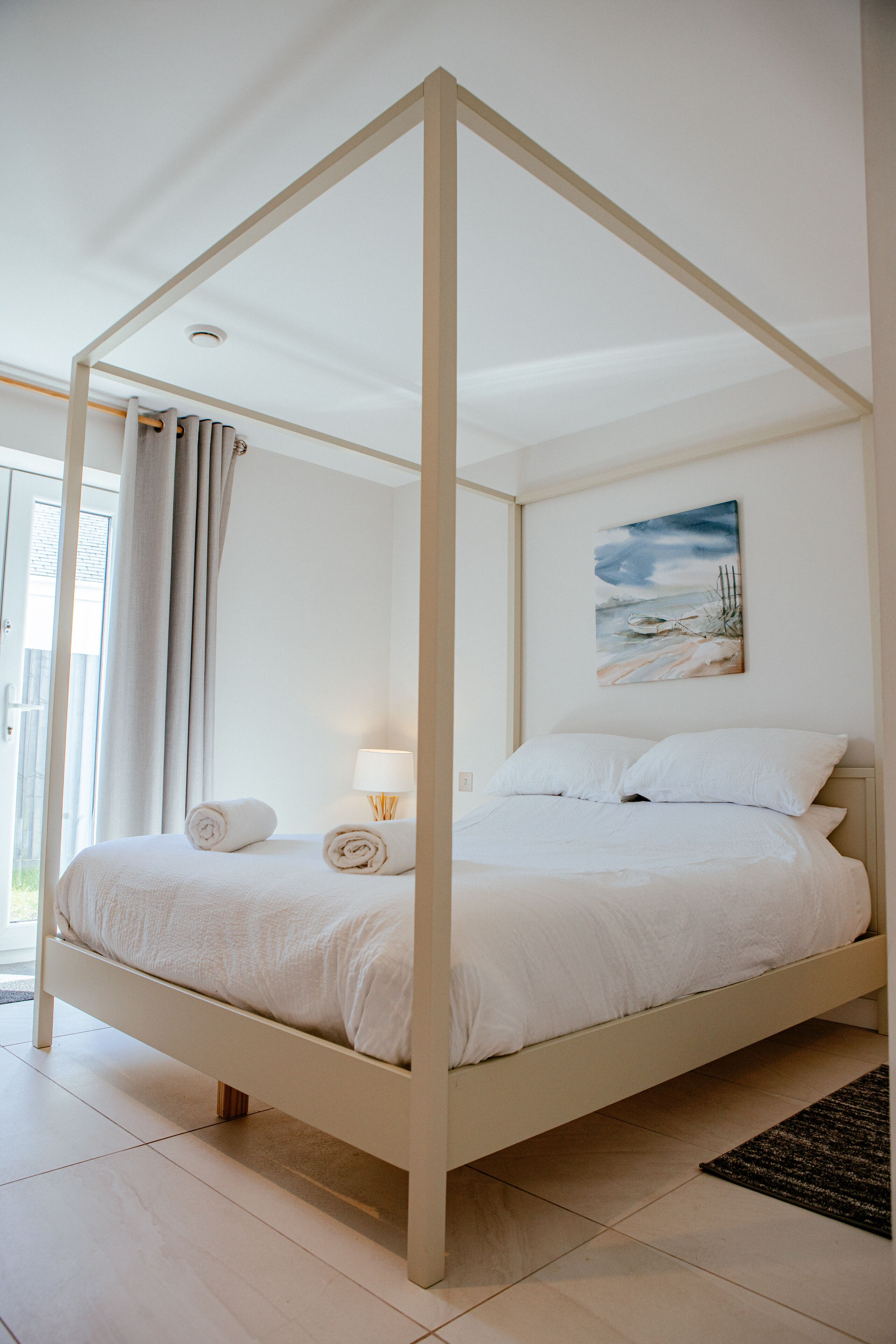 A four-poster bed with white linens, a painting of a beach scene on the wall, and a view of the outdoors through a glass door.