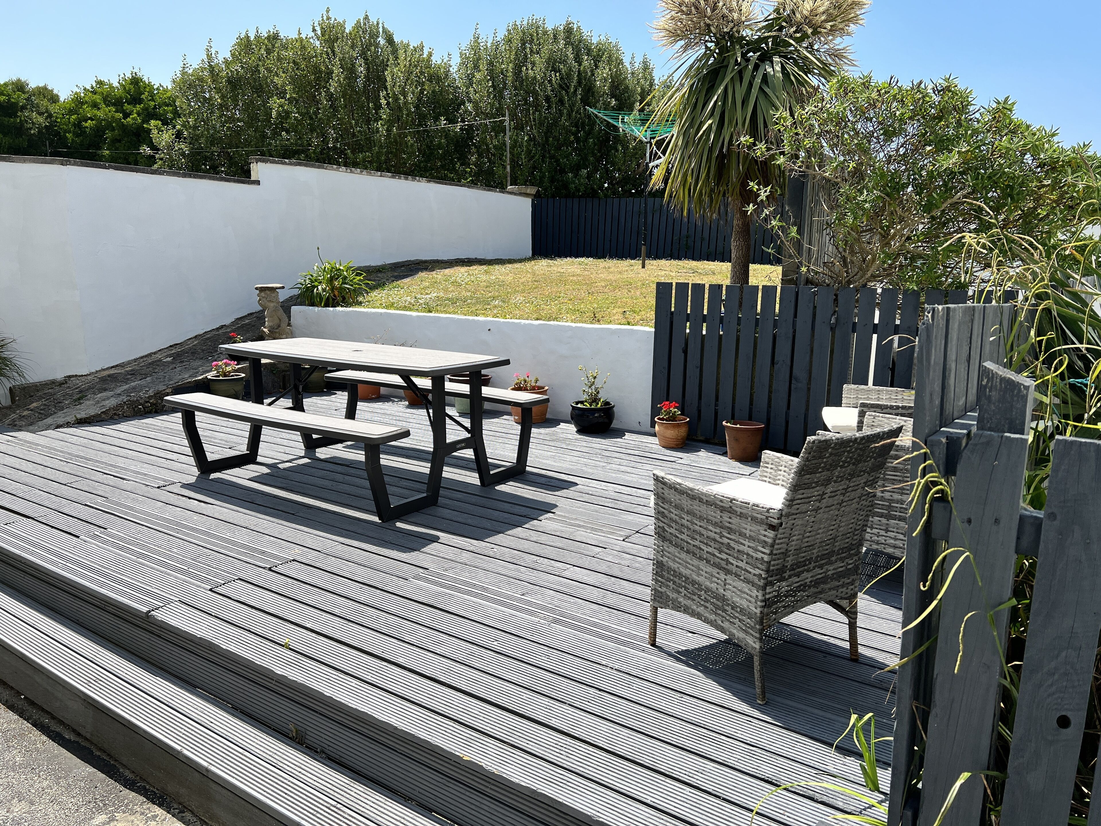 A wooden deck with a picnic table and wicker chairs, surrounded by a low fence and potted plants.
