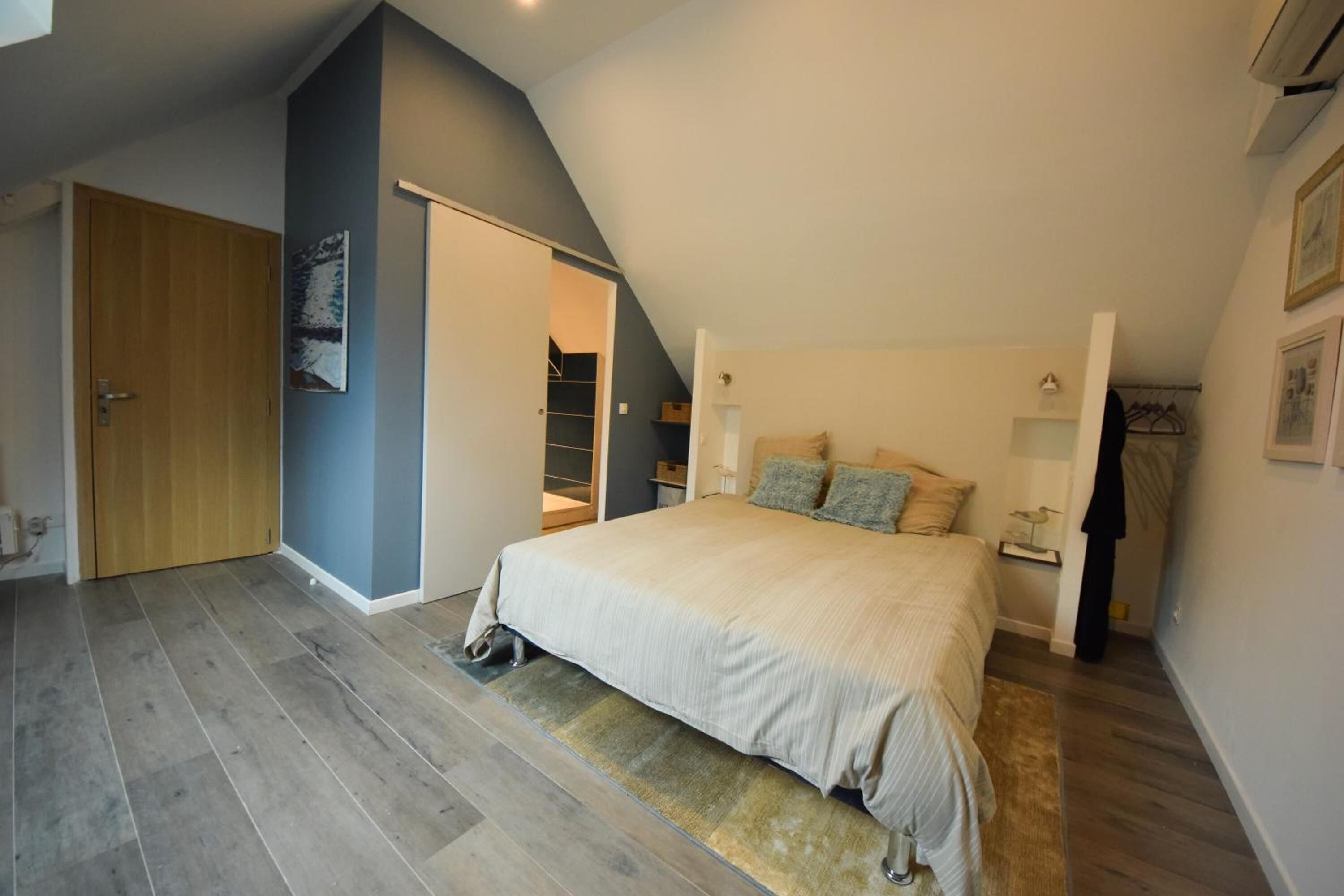 Un dormitorio con techo inclinado, una cama con cubrecama color beige, una puerta de madera y un armario.