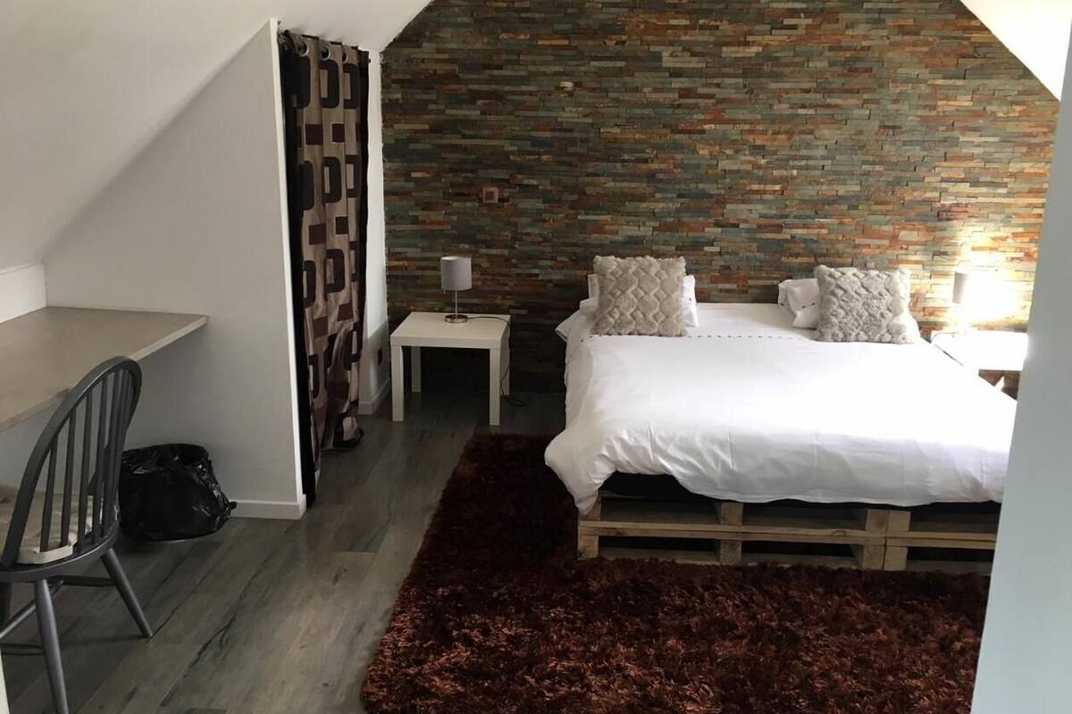 Un dormitorio con techo inclinado, una cama con ropa de cama blanca, una mesita de noche pequeña, una silla y una alfombra marrón oscuro.