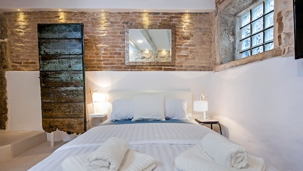 A bedroom with a large bed, two bedside tables, a mirror, and a stone wall. - Luna Rooms and Studios (Rovinj)