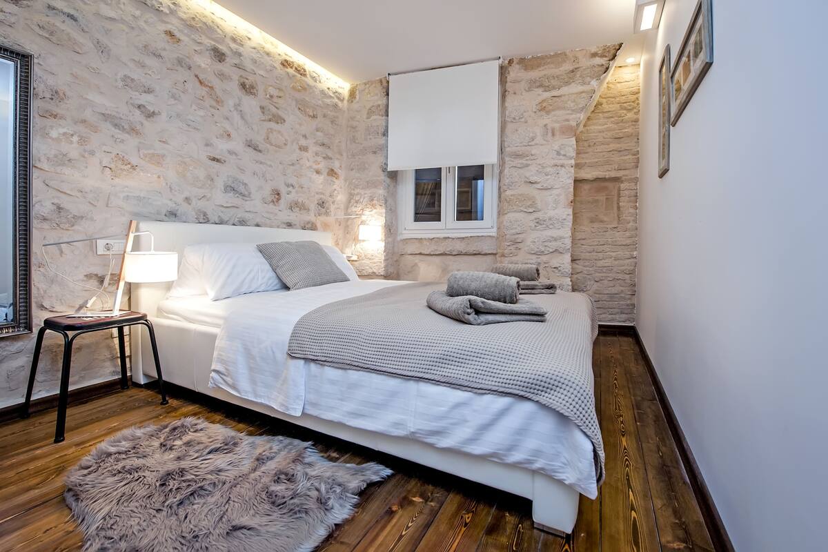 Un dormitorio con pared de piedra, una cama con ropa de cama blanca, una alfombra mullida, una mesita de noche con lámpara y una ventana con persiana blanca.
