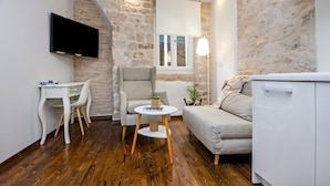 A compact living space with a stone wall, a flat-screen TV, a white desk, a grey armchair, a small round table, a sofa, and a white cabinet. - Luna Rooms and Studios (Rovinj)
