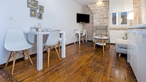 A modern living room with a stone accent wall, a flat-screen TV, and a dining area with white chairs and a small table. - Luna Rooms and Studios (Rovinj)