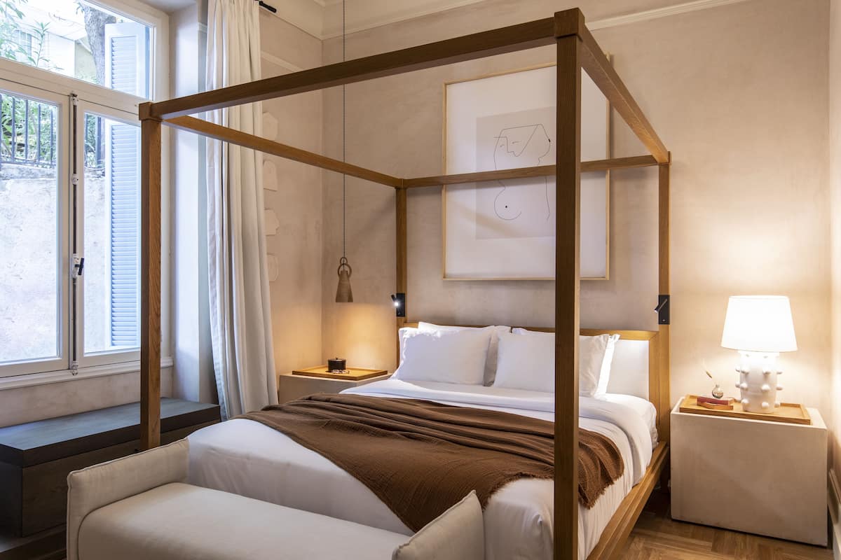 A four-poster bed with a wooden frame, two bedside tables with lamps, a window with blinds, and a framed artwork on the wall.
