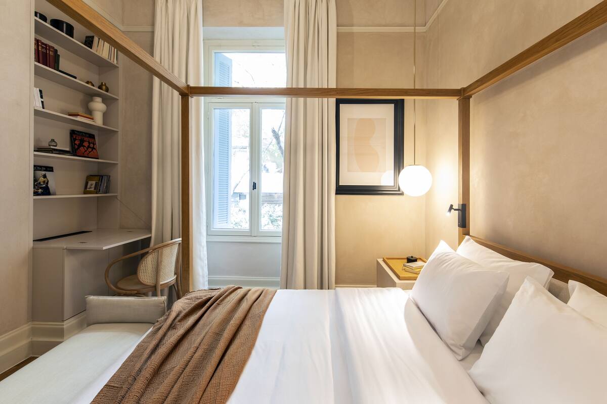 A hotel room with a bed, a desk, a chair, a bookshelf, and a window with curtains.