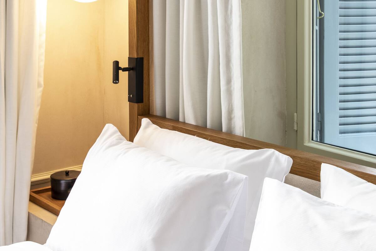 A neatly made bed with white linens, a bedside lamp, and a window with blinds.
