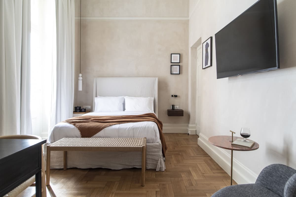 A hotel room with a bed, a small table with a glass of wine, a television mounted on the wall, and a wooden floor.