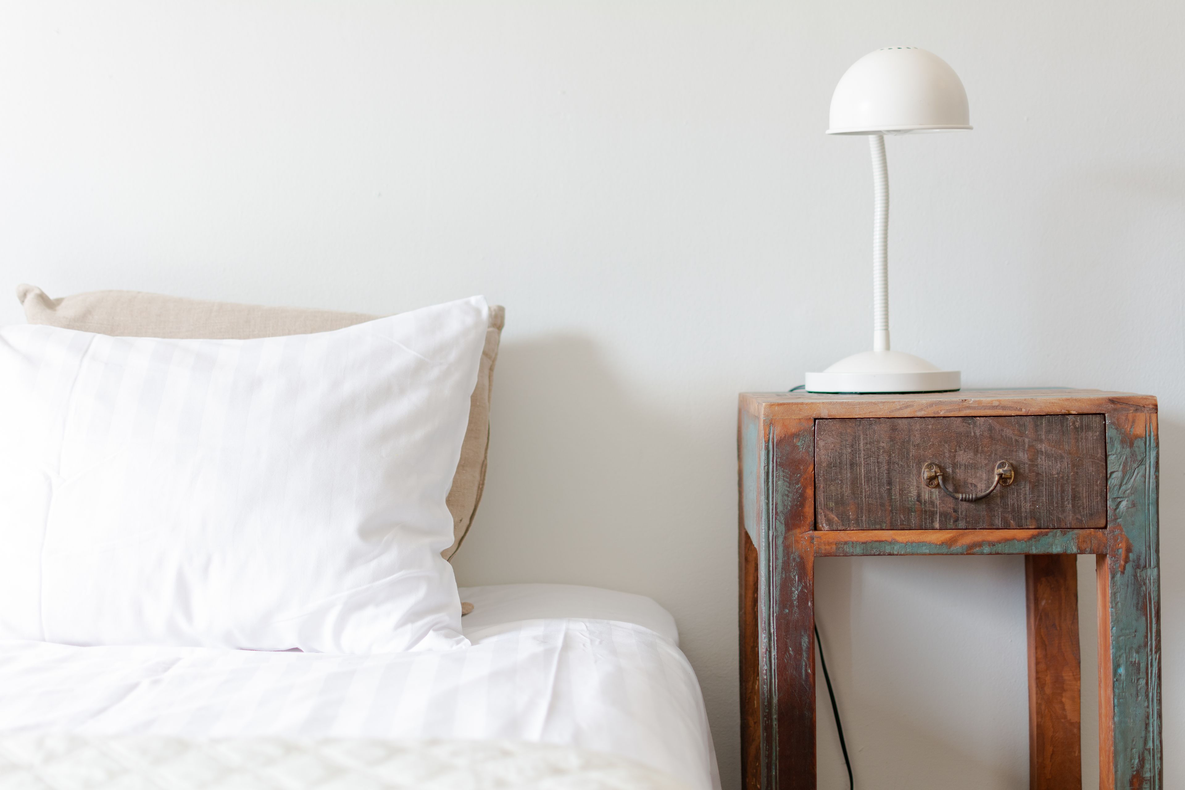 Ein Bett mit weißer Bettwäsche, einem weißen Kissen und einer weißen Lampe auf einem Nachttisch aus Holz mit einer Schublade.