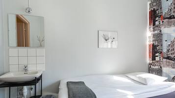 A bedroom with a white bed, a bathroom with a sink and mirror, and a wall with a framed picture.