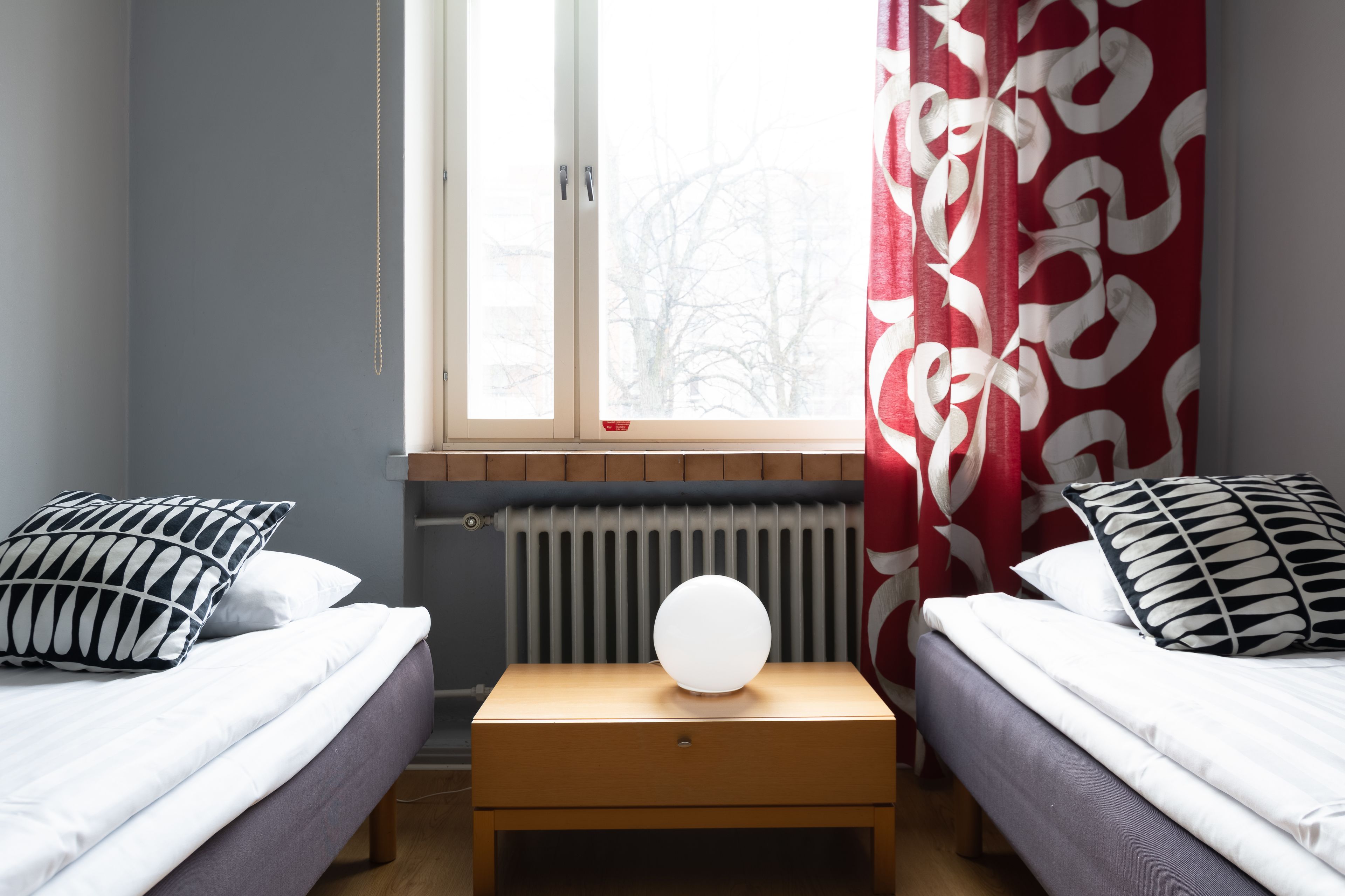 A bedroom with two beds, a bedside table, a radiator, and a window with curtains.