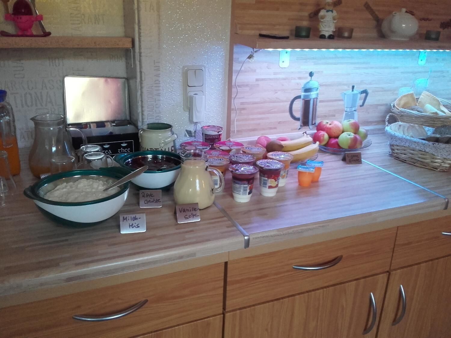 A kitchen counter with various food items and containers, including jars of preserves, a bowl of oatmeal, and a pitcher of milk.