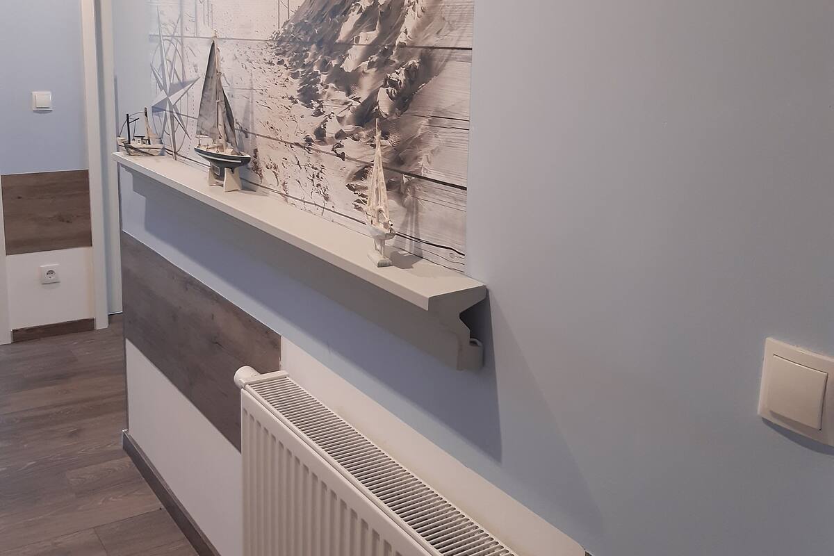 A hallway with a wooden floor, a white radiator, and a wall-mounted shelf with a model ship and a painting.