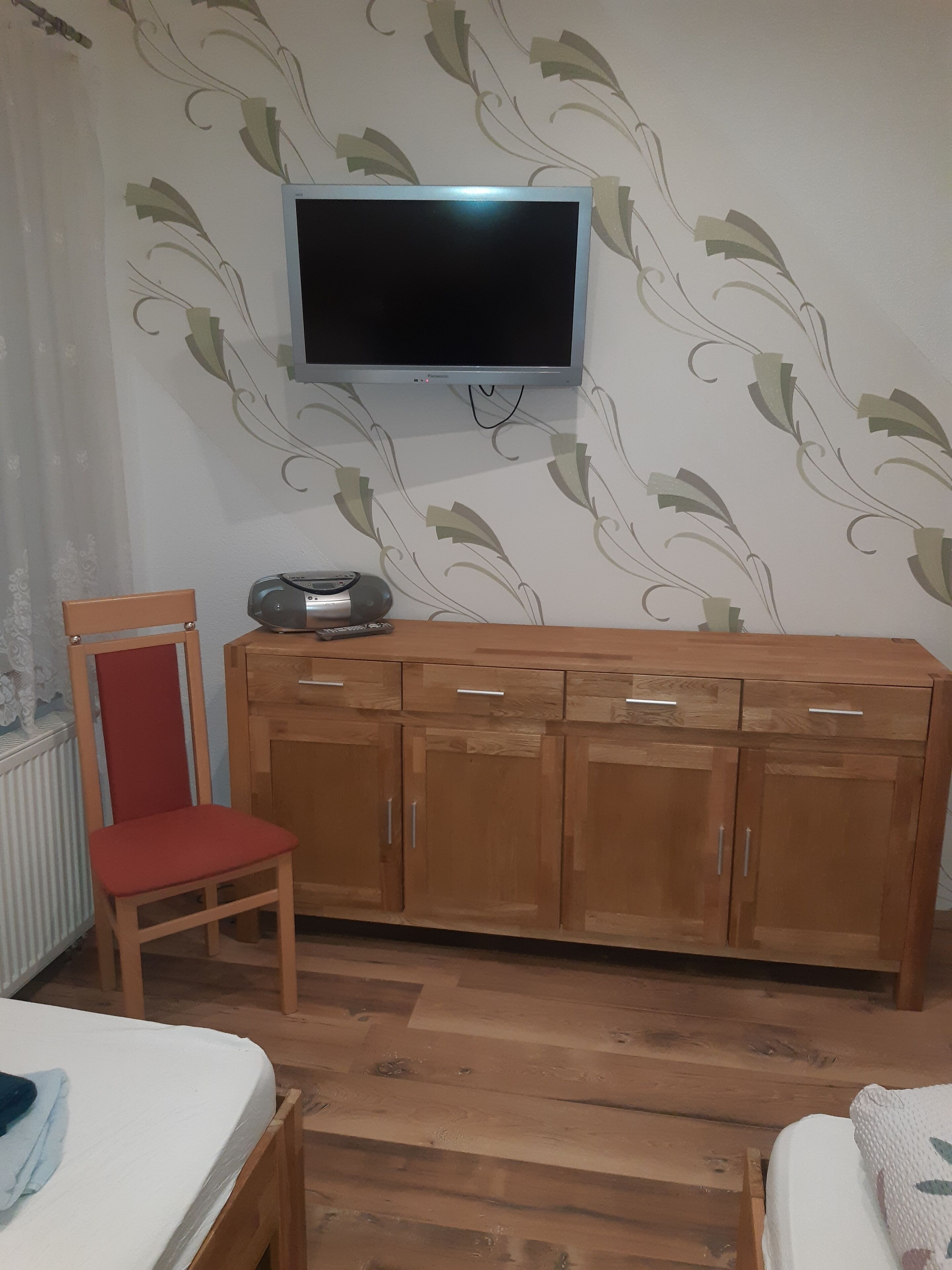 Ein Zimmer mit einem Holzkasten, einem an der Wand befestigten Fernseher, einem roten Sessel und einem Bett mit weißer Matratze.