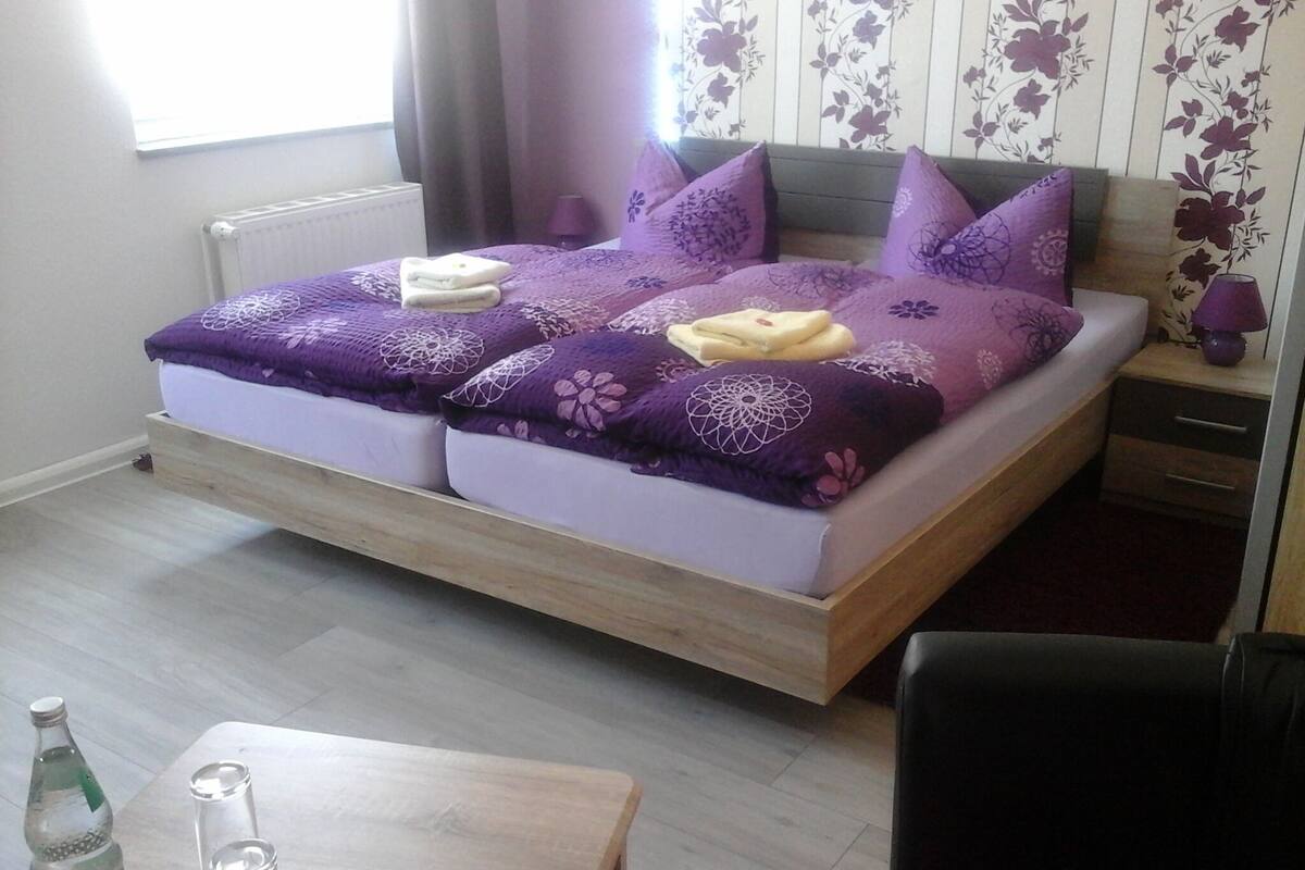 A double bed with purple bedding, a bedside table with a lamp, and a small table with bottles and glasses.