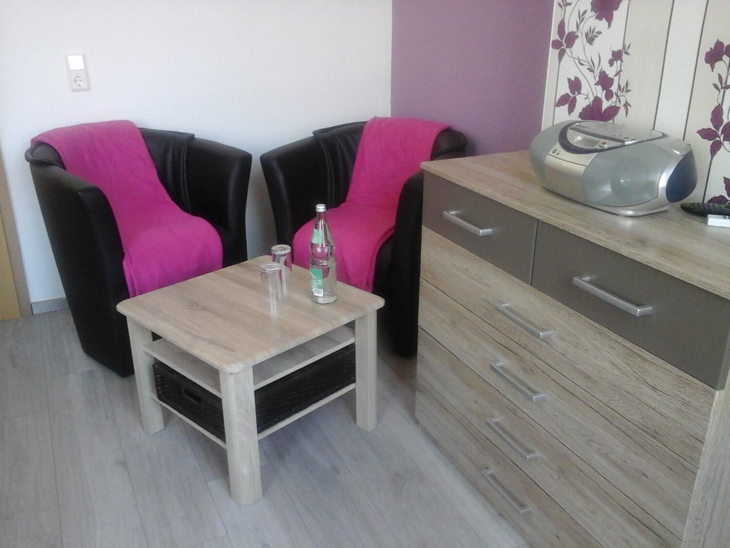 A small kitchenette with a wooden table, two black chairs with pink cushions, a microwave, and a cabinet with drawers.