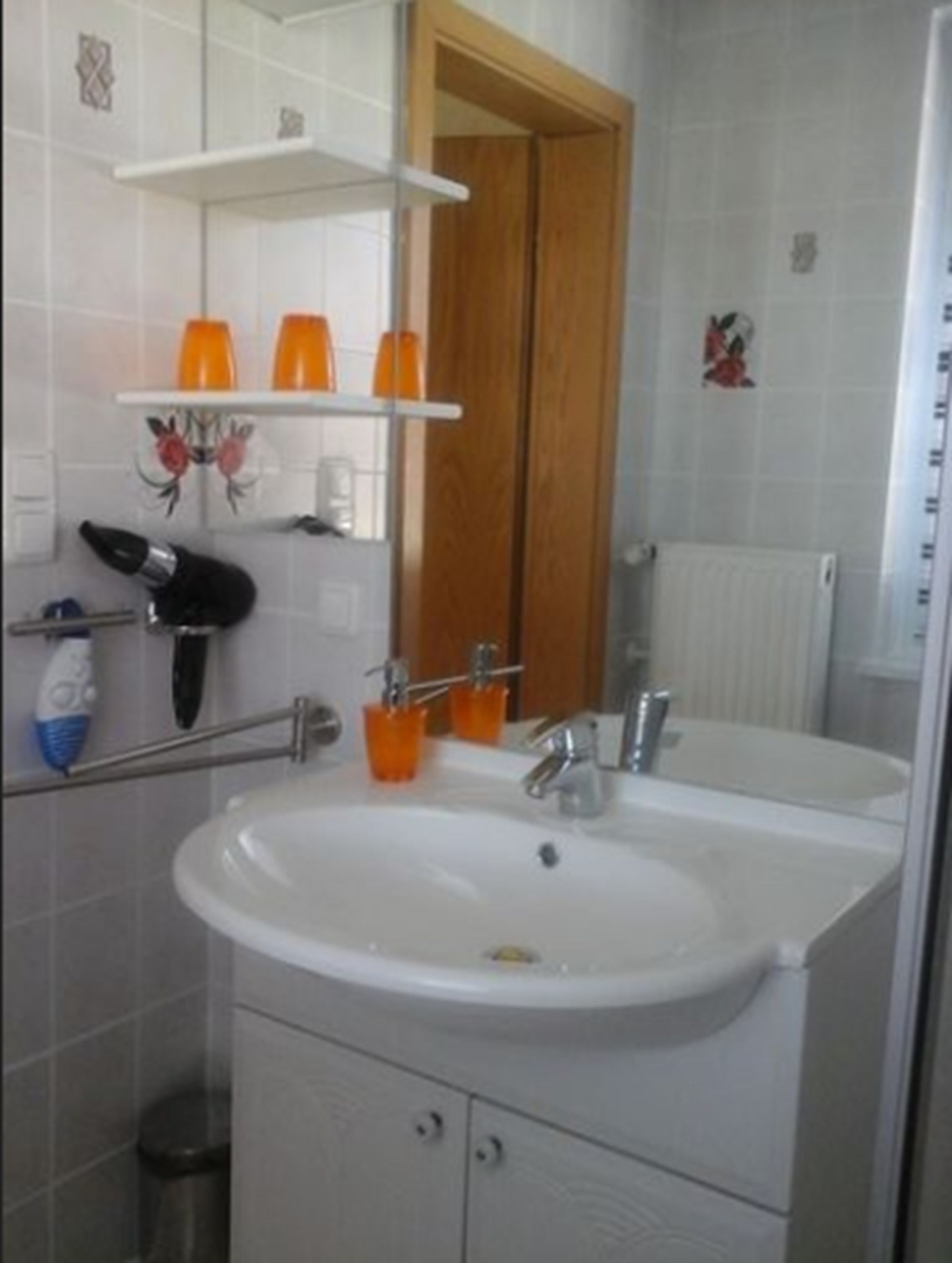 Un bany amb un lavabo blanc, gots taronja sobre una estantalla, un mirall i una porta de fusta.