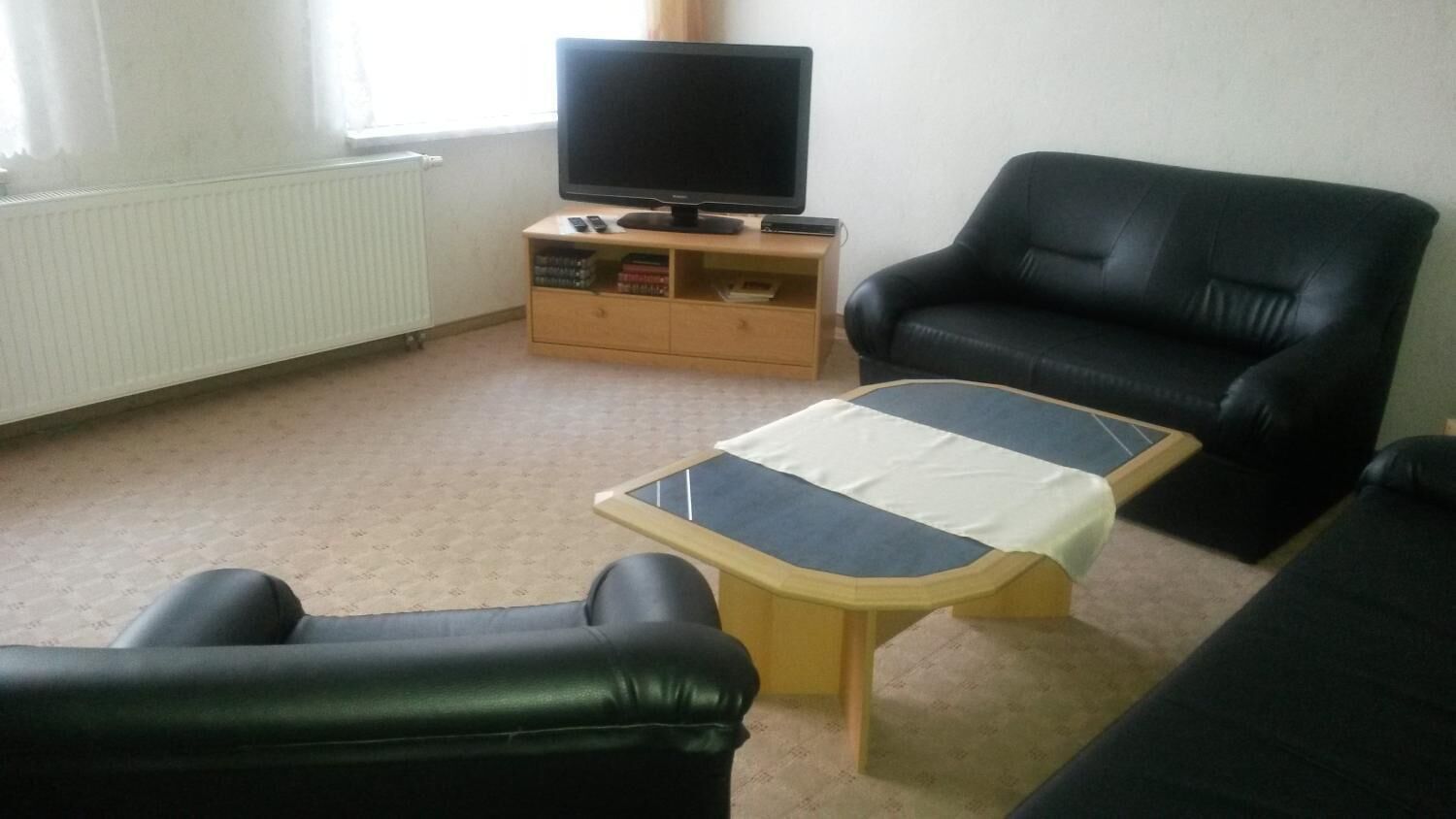 A living room with a black leather sofa, a wooden coffee table, a television on a wooden stand, and a radiator.