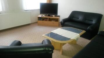 A living room with a black leather sofa, a wooden coffee table, a television on a wooden stand, and a radiator.