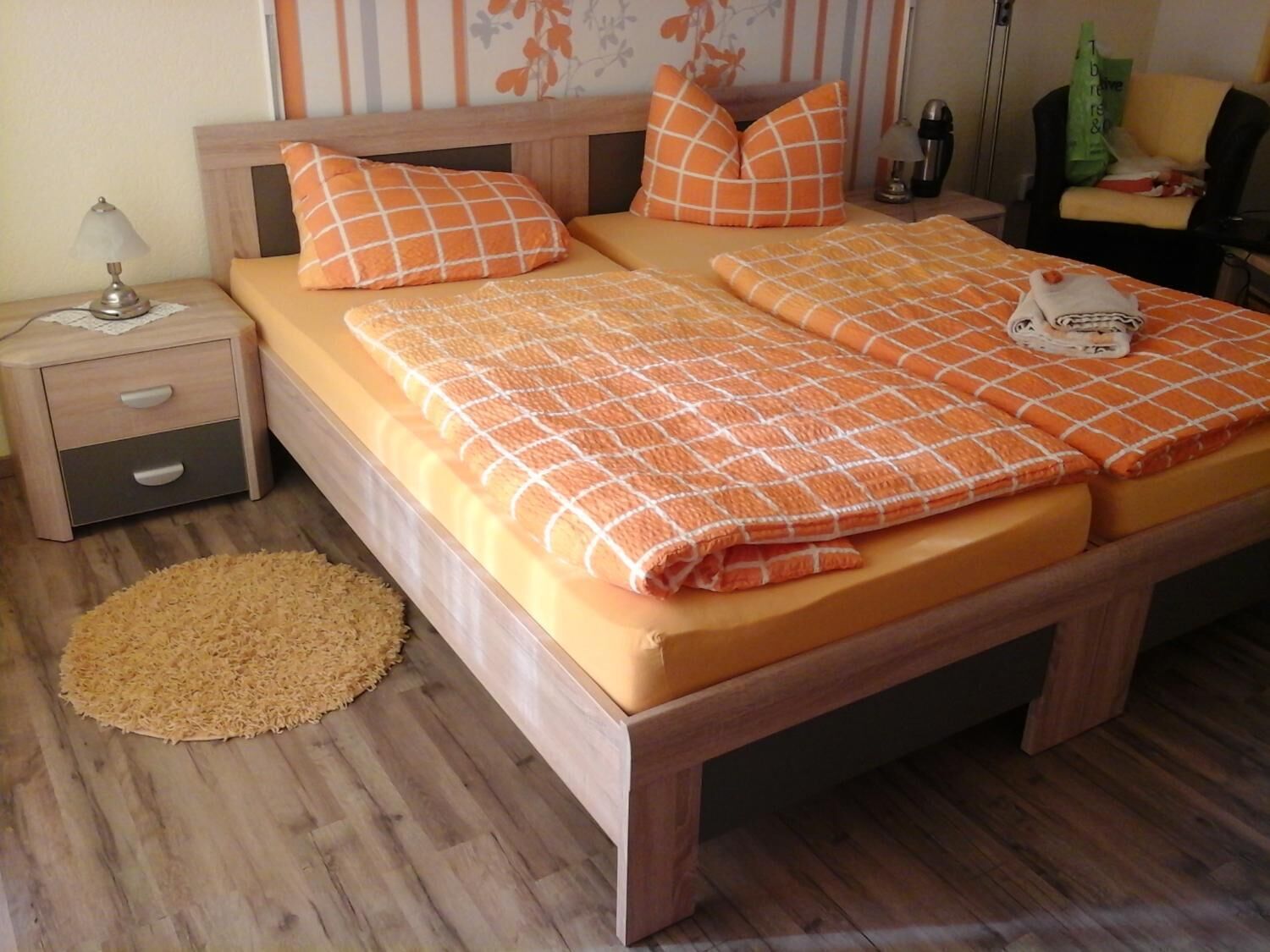 A double bed with orange bedding, a bedside table with a lamp, and a small rug on a wooden floor.
