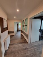 Um corredor com piso de madeira, paredes brancas e um radiador. Há portas em ambos os lados e uma janela no final do corredor.
