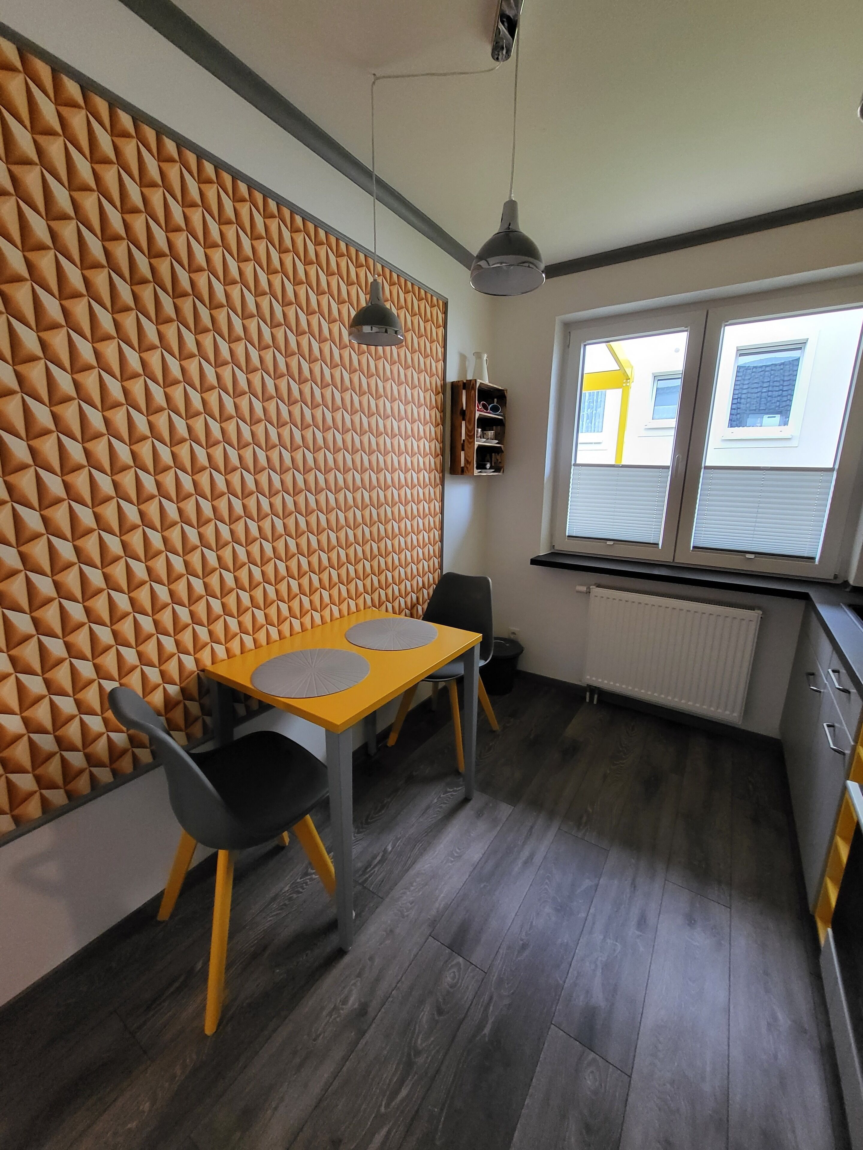 A modern kitchen with a geometric patterned wall, a dining area with a yellow table and black chairs, and a window with a view of a yellow building.