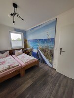 Quarto com uma janela grande, cama com roupa de cama floral, ventoinha de teto e um mural com cena de praia.