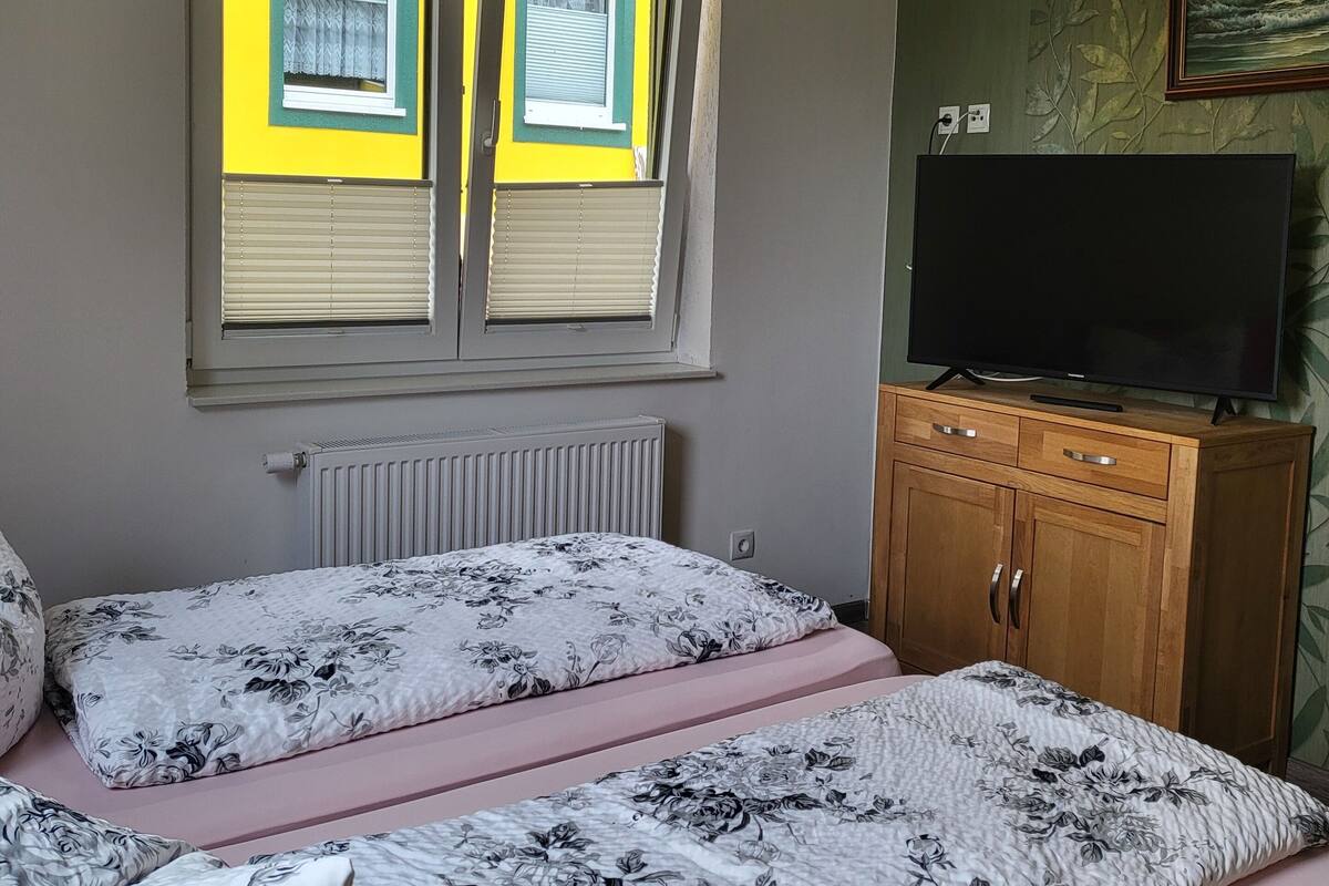 A bedroom with two beds, a radiator, a wooden cabinet with a television, and a window with blinds.