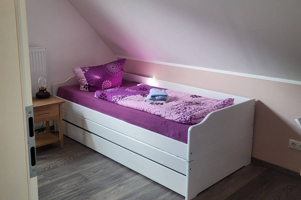 A bedroom with a white bed, purple bedding, a nightstand, and a wooden floor.