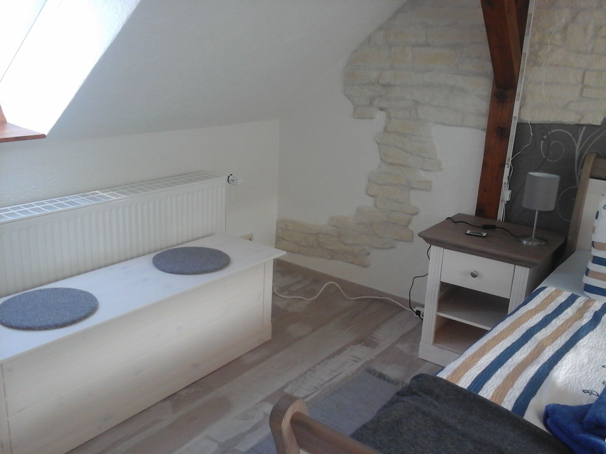 Una stanza con soffitto spiovente, parete in pietra, comodino bianco con lampada, letto con biancheria a righe e cuscino rotondo su una panca.