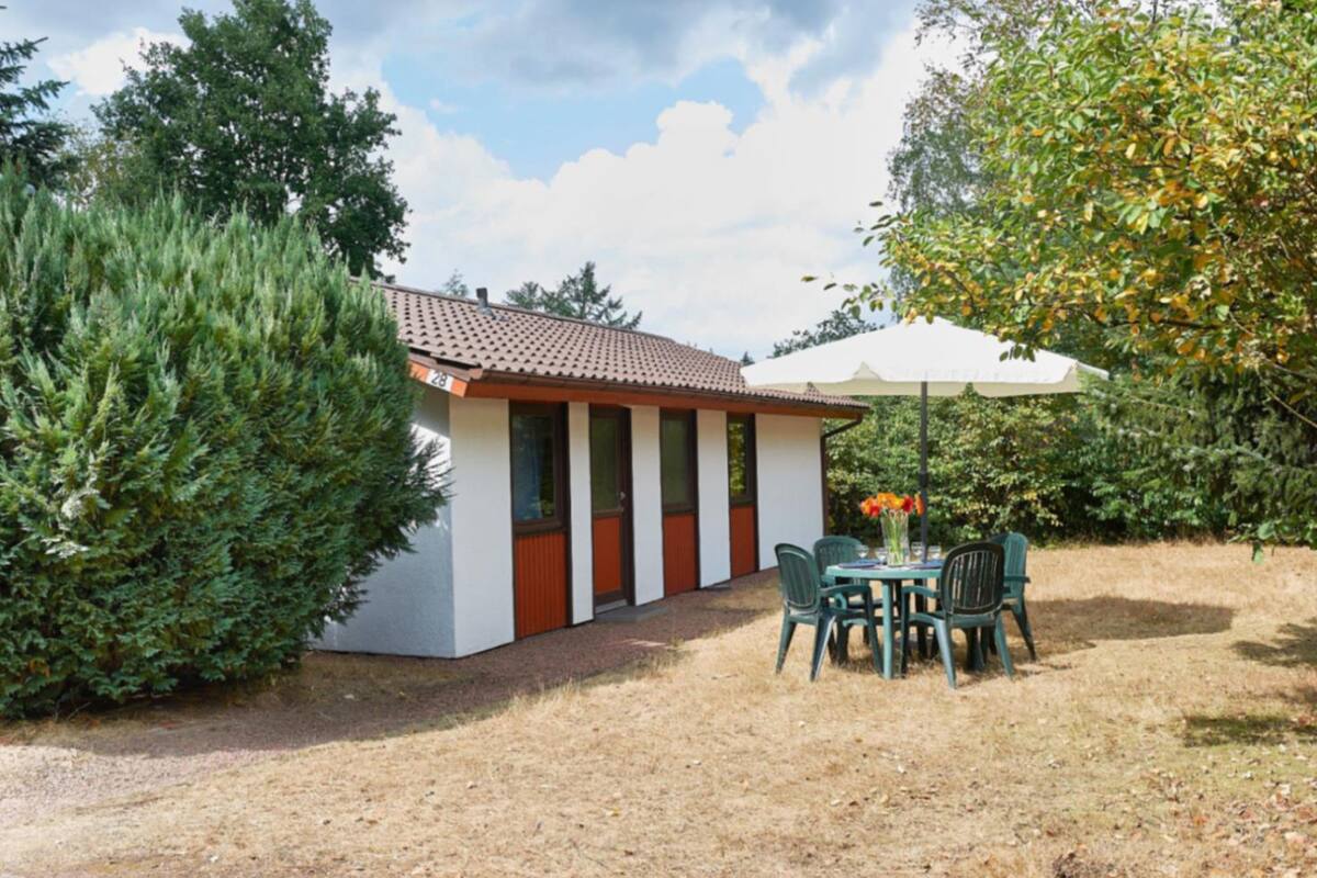 Un petit bâtiment avec un toit en tuiles, un espace extérieur abrité avec des chaises et une table, et un grand arbre sur la droite.