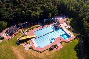 An aerial view of a swimming pool complex with a slide, surrounded by greenery and a parking area.