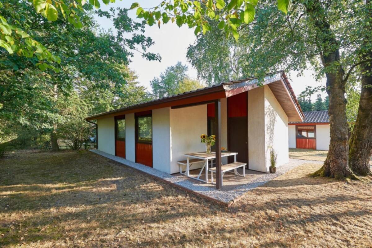 Un bâtiment moderne, de plain-pied, doté d’un toit en pente, d’un patio couvert avec une table et des chaises, et d’un petit jardin.