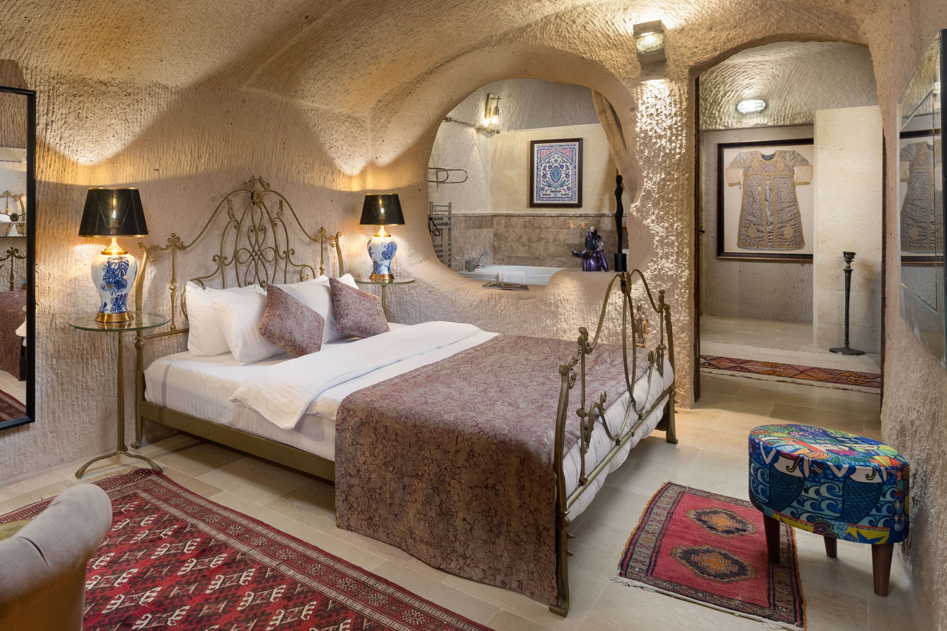 A bedroom with a bed, bedside tables, a nightstand, a mirror, a wall-mounted shelf, and a small stool.