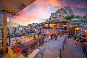 A rooftop terrace with outdoor seating, a view of a mountainous landscape, and a building with a wooden facade. - The Rupestral House Hotel (Nevsehir)