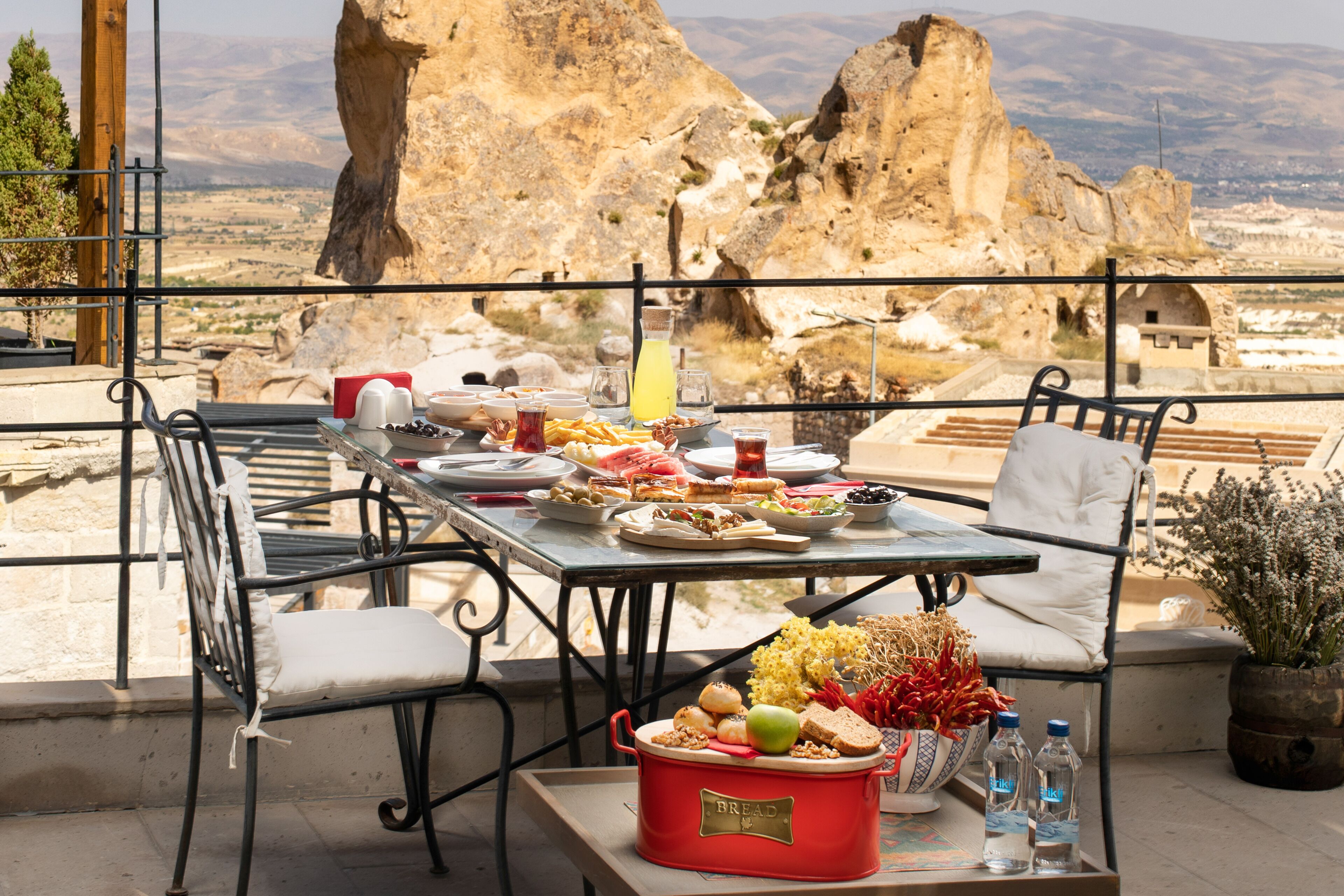 Mesa de comedor al aire libre con diferentes platos, una fuente roja para pan y vistas a terreno rocoso.