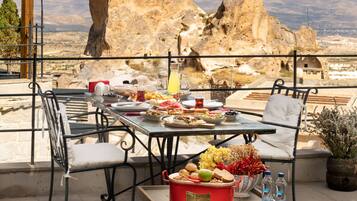 Mesa de comedor al aire libre con diferentes platos, una fuente roja para pan y vistas a terreno rocoso.