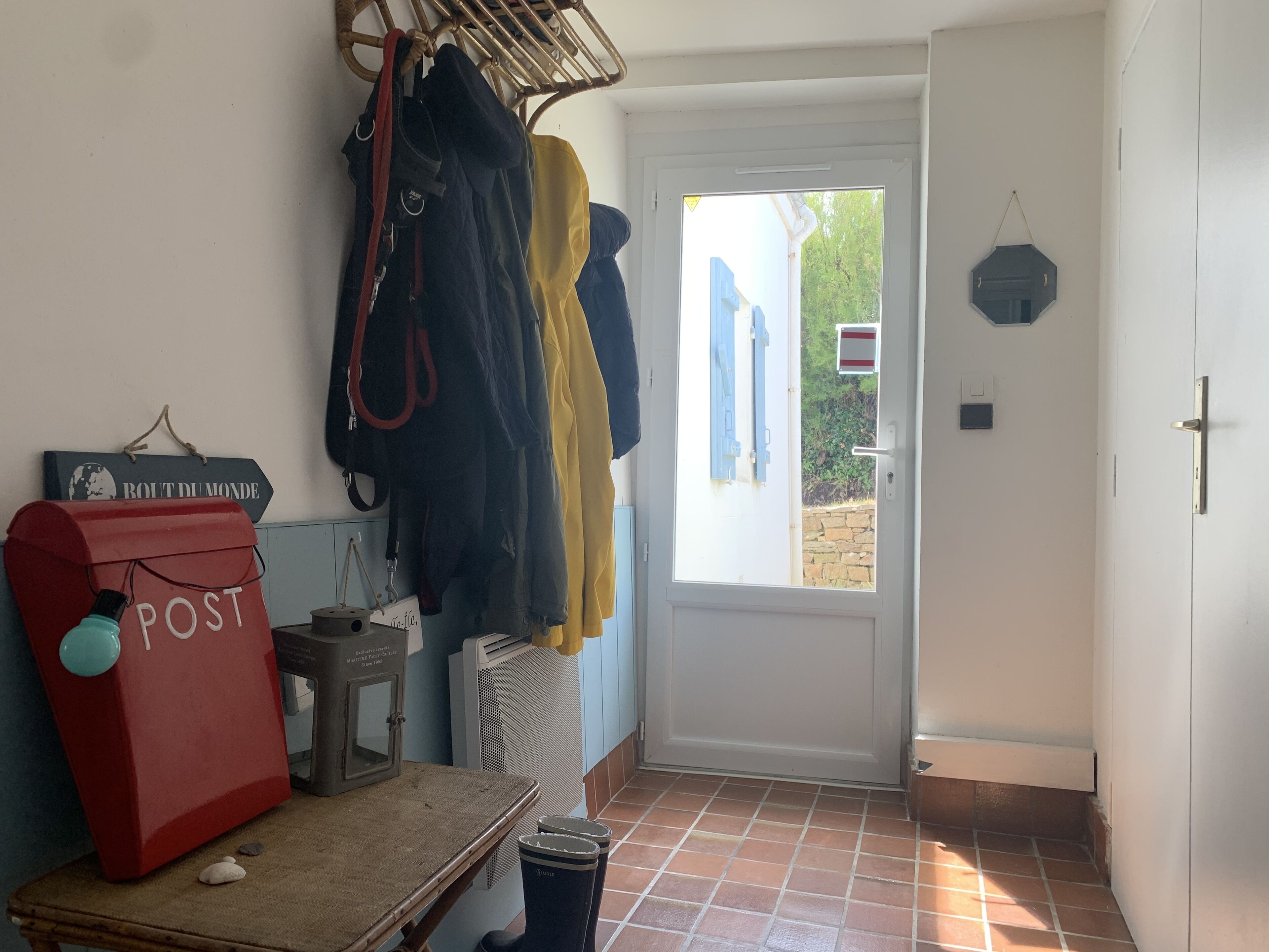 Un couloir avec une boîte aux lettres rouge portant l’inscription « POSTE », un étendoir à linge, une petite table avec une bouilloire et une porte donnant sur l’extérieur.