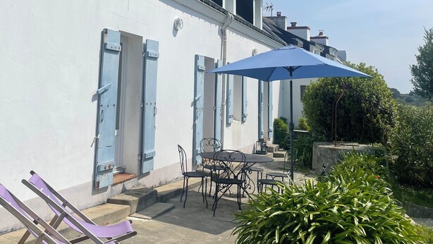 Un patio con una mesa y sillas bajo un paraguas azul, un edificio blanco con contraventanas azules y un pequeño jardín con una planta.