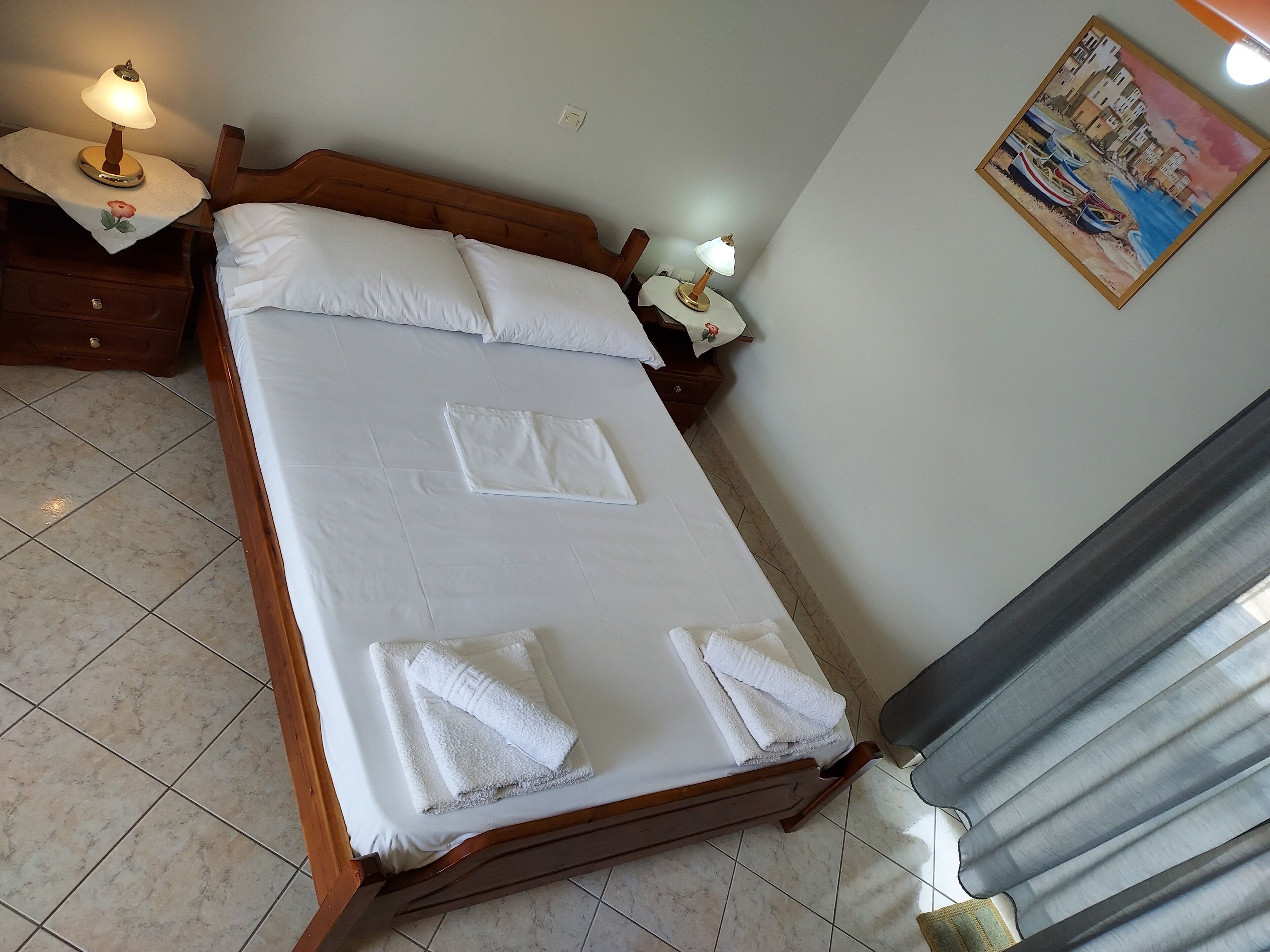 Una cama bien hecha con sábanas y toallas blancas, cabecera de madera, mesita de noche con lámpara y un cuadro en la pared.