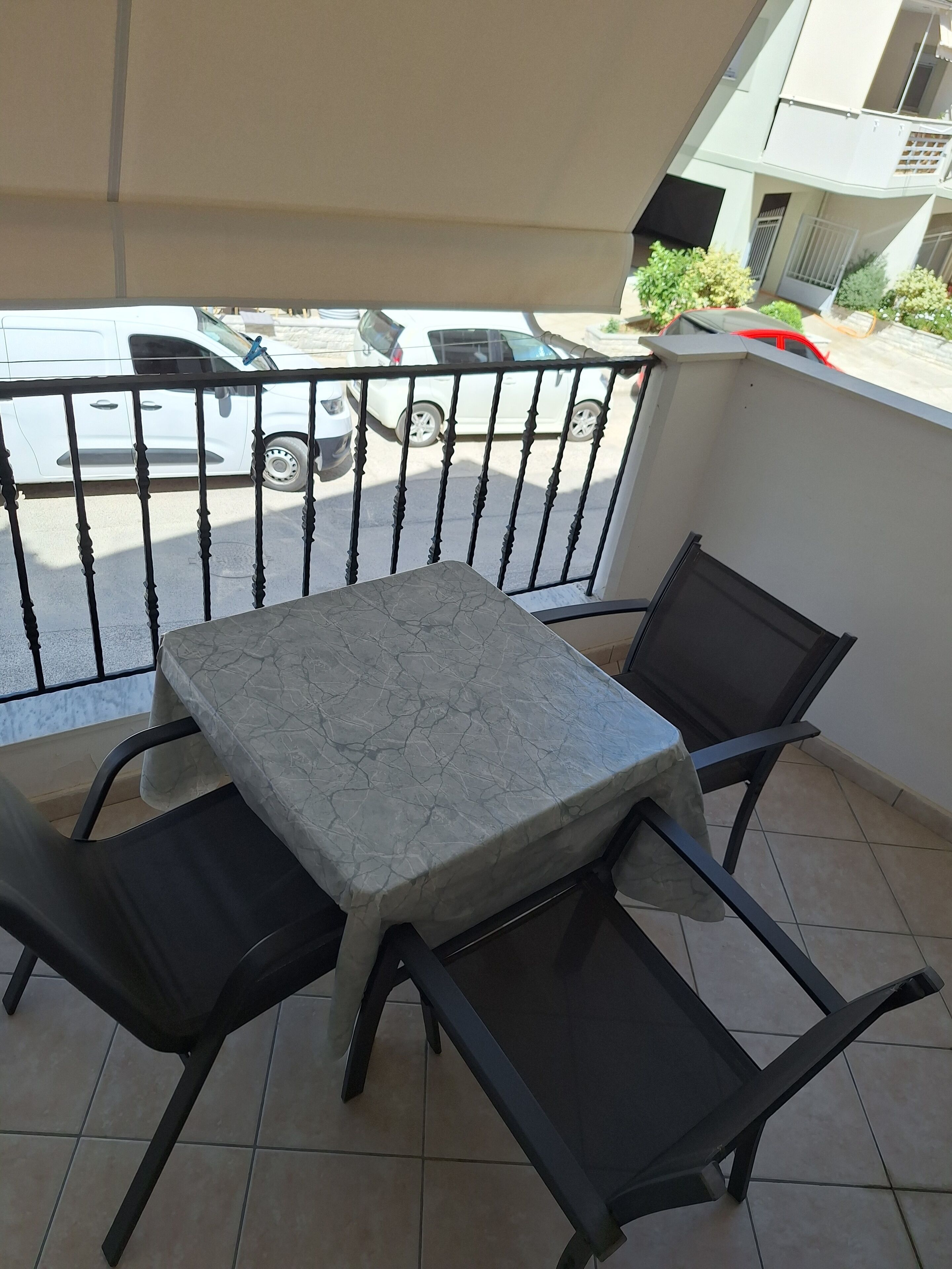 A balcony with a table and chairs, a view of parked cars, and a building in the background.