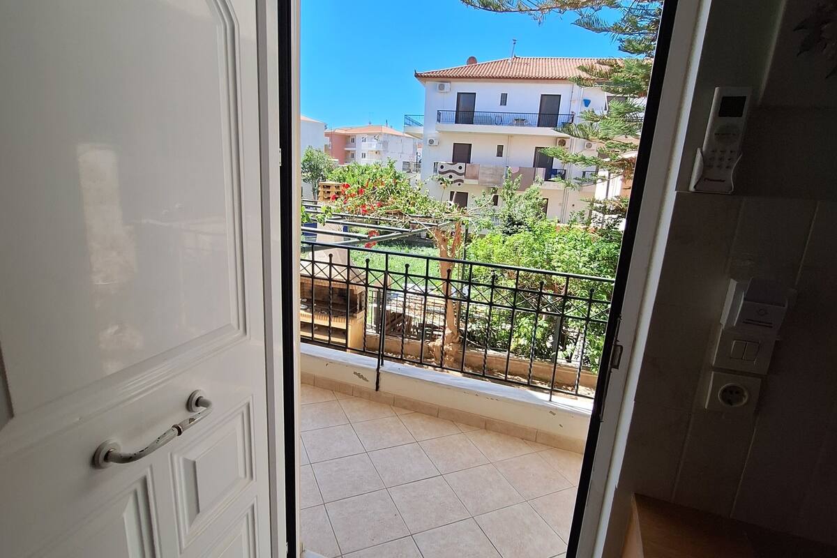 A view from a balcony with a tiled floor, a white door, and a visible outdoor area with buildings and greenery.