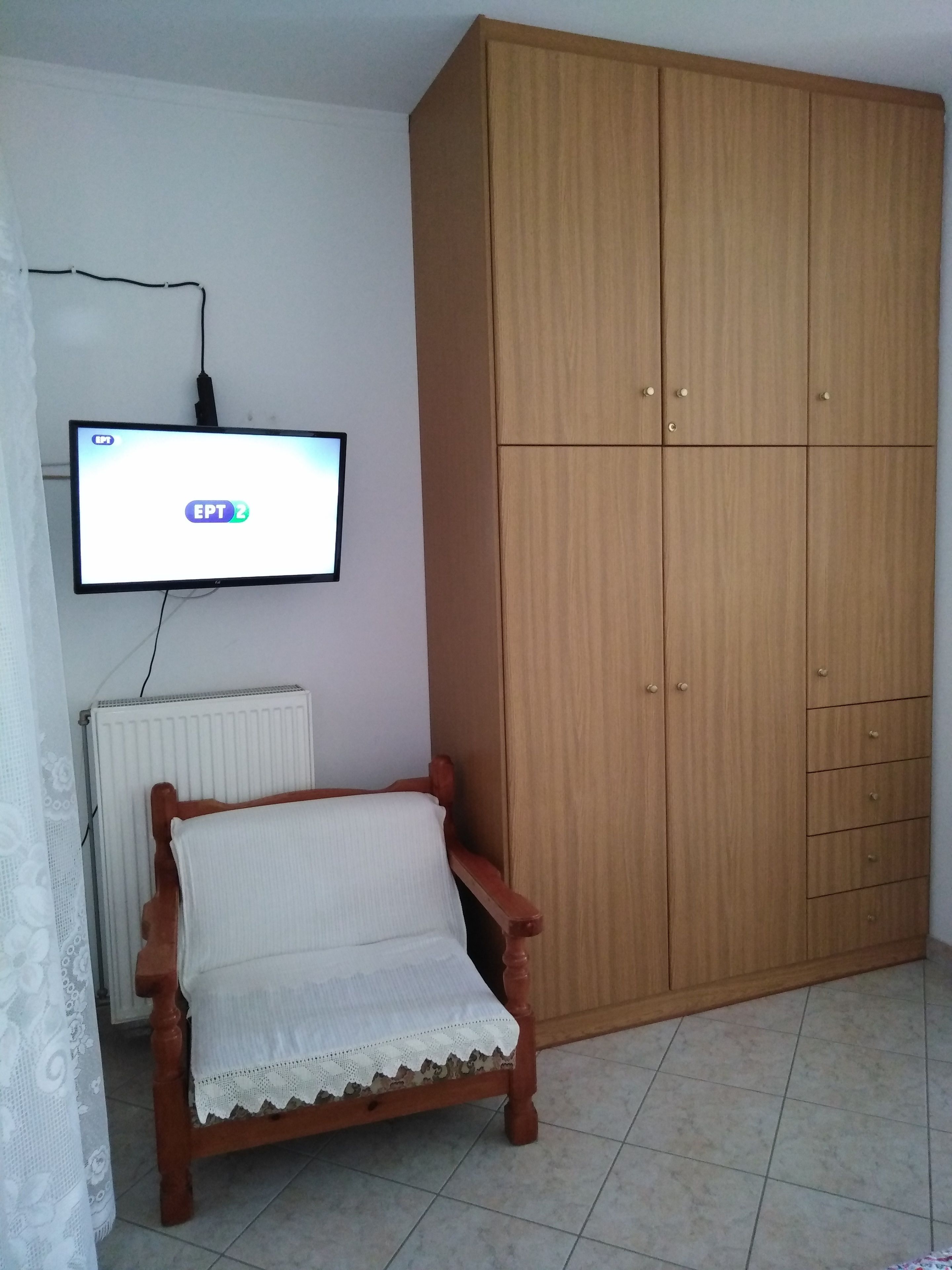 A room with a wooden wardrobe, a wooden chair with a white cushion, a television mounted on the wall, and a radiator.