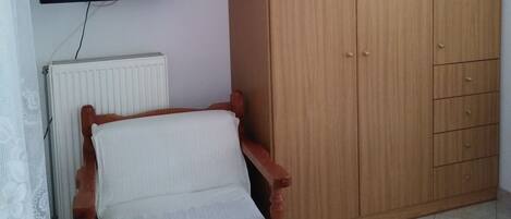 A room with a wooden wardrobe, a wooden chair with a white cushion, a television mounted on the wall, and a radiator.