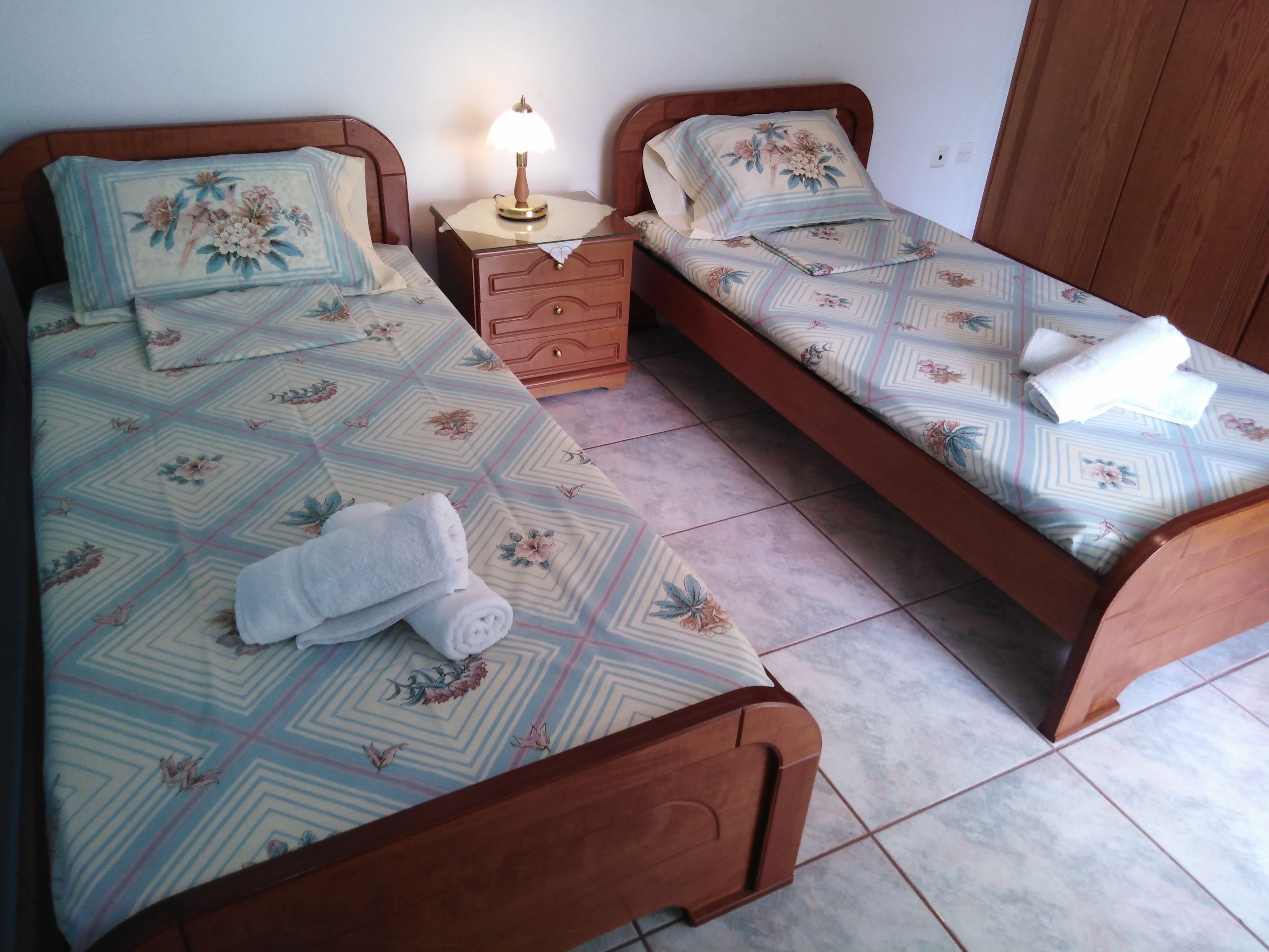 A bedroom with two single beds, each with floral-patterned bedding and a wooden headboard. There is a nightstand with a lamp between the beds, and a wooden dresser to the side.