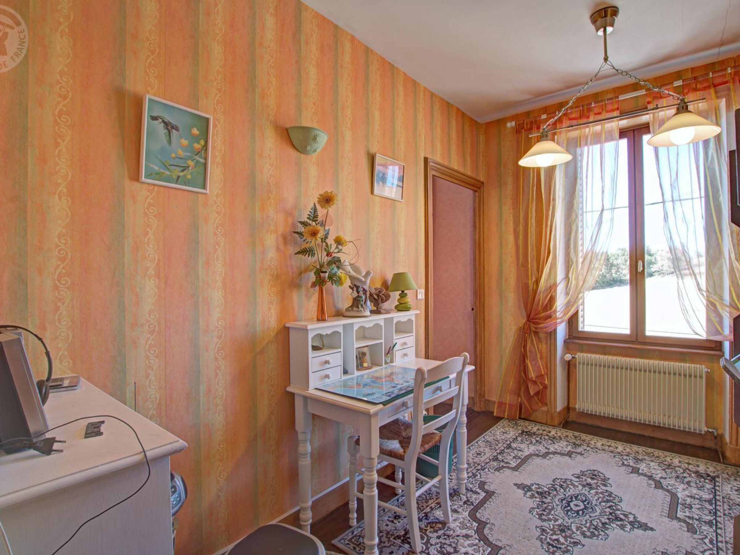 A room with striped wallpaper, a desk with a computer, a chair, a radiator, and a window with curtains.